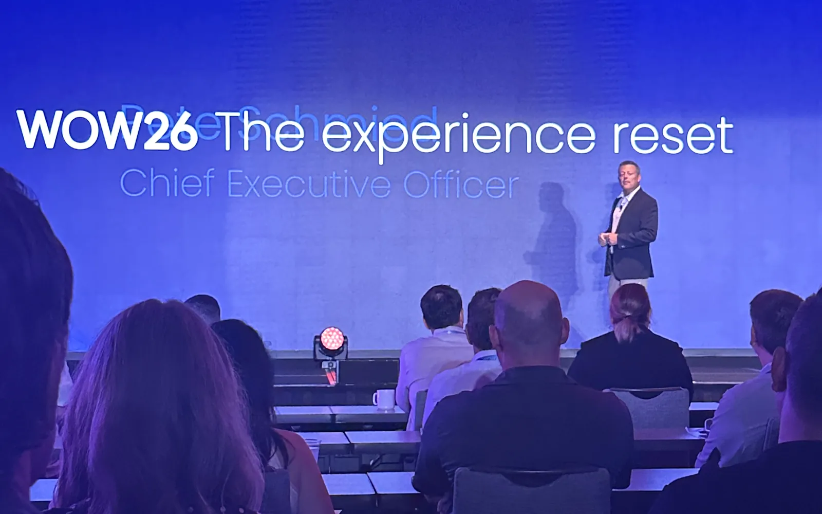 Appspace CEO Pete Schmied speaking on the WOW26 stage in front of a screen that reads WOW26, The experience reset, with attendees seated in the foreground.
